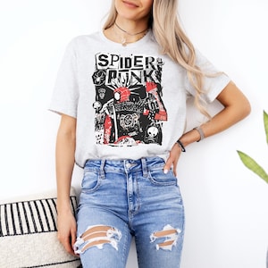 May include: A light gray t-shirt with the words "SPIDER PUNK" in a black, red, and white graphic design. The graphic features a stylized illustration of a character. The shirt is paired with blue jeans.