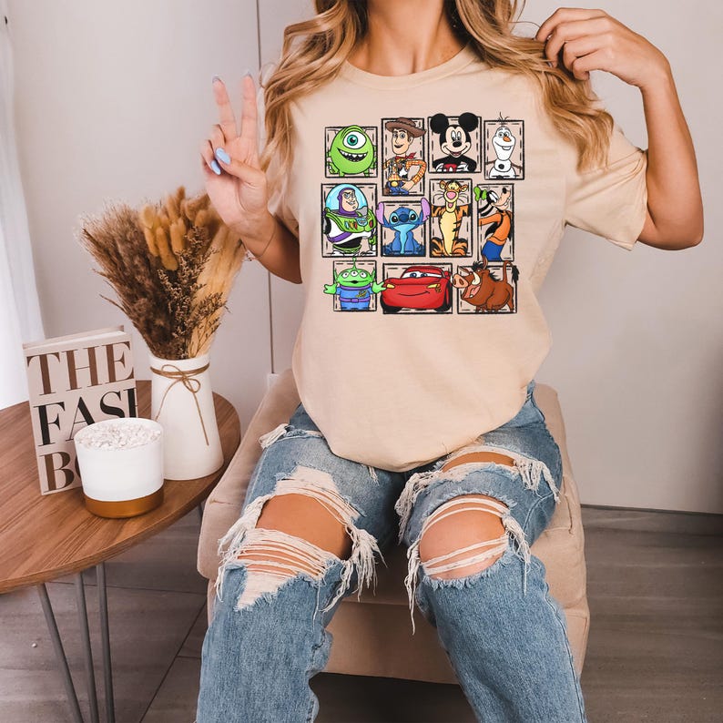 May include: Beige t-shirt featuring a grid of cartoon characters including Mickey Mouse, Buzz Lightyear, and Stitch. The shirt has a crew neck and short sleeves. The design is printed in vibrant colors on the front of the shirt.