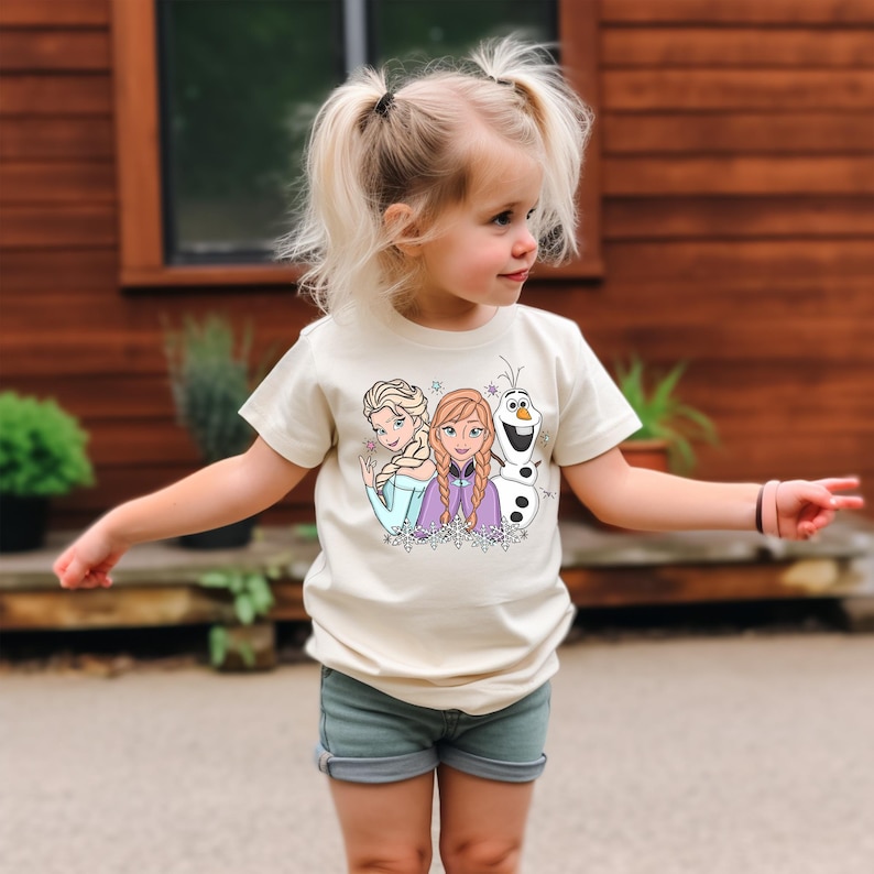 Puede incluir: Una ni&ntilde;a peque&ntilde;a con una camiseta color crema con un gr&aacute;fico de Elsa, Anna y Olaf de la pel&iacute;cula Frozen. La ni&ntilde;a tambi&eacute;n lleva pantalones cortos vaqueros. El fondo es una pared de madera.