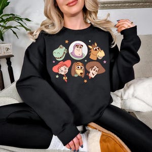 May include: Black crewneck sweatshirt featuring cartoon character portraits from a popular animated film. The design includes images of a green dinosaur, Buzz Lightyear, a horse, Jessie, Slinky Dog, Woody, and other characters.