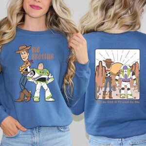 Op de afbeelding: Een blauwe sweatshirt met de tekenfilmfiguren Woody en Buzz Lightyear. De voorkant van de sweatshirt heeft de woorden "Big Brother" boven de karakters. De achterkant van de sweatshirt heeft een woestijnsc&egrave;ne met de woorden "You've Got A Friend In Me".