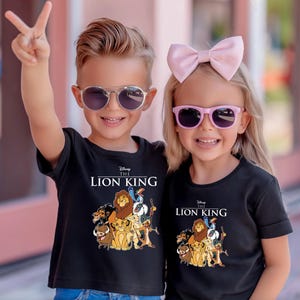 May include: Two children wearing black t-shirts with "The Lion King" graphic and characters. The boy wears silver sunglasses and the girl wears pink sunglasses and a pink bow. The boy is holding up two fingers.