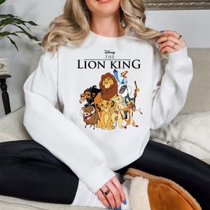 May include: White sweatshirt featuring a colorful graphic of characters from Disney's The Lion King. The text "Disney The Lion King" is printed above the graphic. The sweatshirt is a crew neck style.