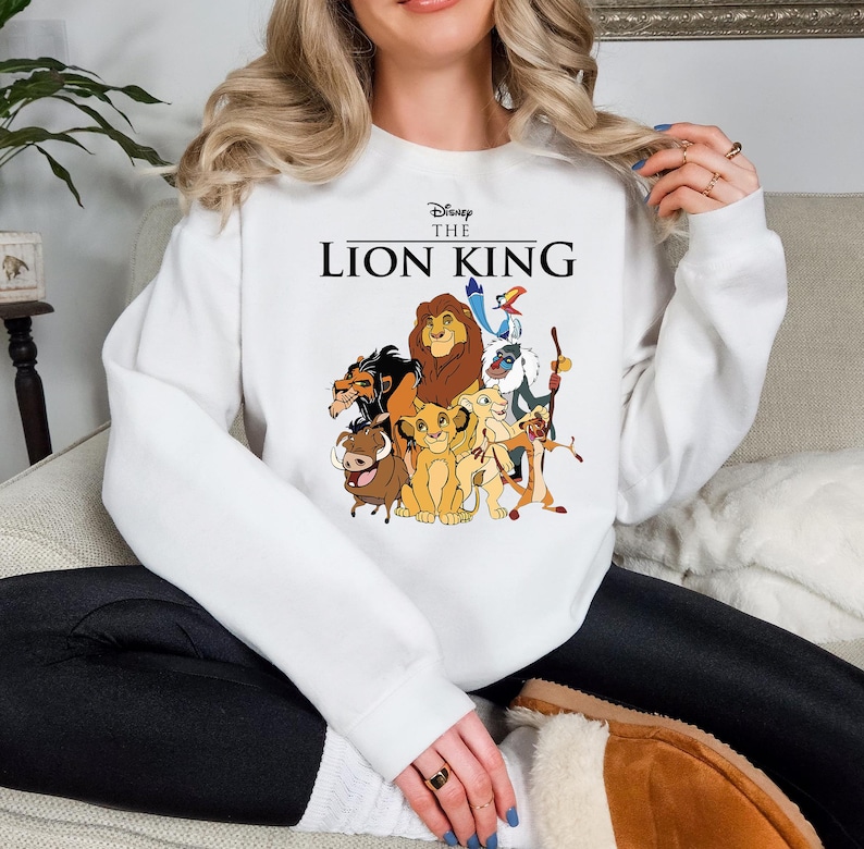 May include: White sweatshirt featuring a colorful graphic of characters from Disney's The Lion King. The text "Disney The Lion King" is printed above the graphic. The sweatshirt is a crew neck style.
