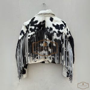 May include: A cropped jacket made of black and white cowhide featuring a pointed collar and long fringe detailing along the back and sleeves. The jacket displays a natural animal print pattern and includes a brown logo overlay with the text COWHIDE.