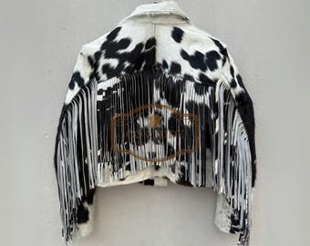 May include: A cropped jacket made of black and white cowhide featuring a pointed collar and long fringe detailing along the back and sleeves. The jacket displays a natural animal print pattern and includes a brown logo overlay with the text COWHIDE.