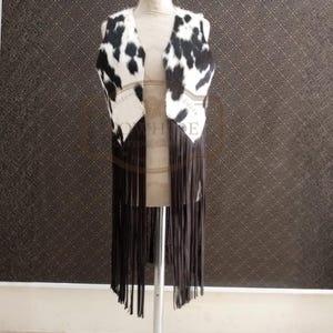 May include: A sleeveless vest with a cowhide pattern in black and white. The vest has long, dark brown fringe along the bottom. The vest is displayed on a mannequin.