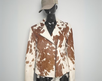 May include: A brown and white cowhide leather jacket with a zipper closure. The jacket has a  asymmetrical zipper and is on a mannequin wearing a grey baseball cap.