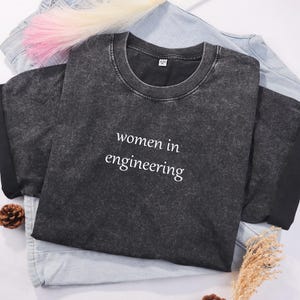 May include: A charcoal grey t-shirt with the text "women in engineering" in white. The shirt is folded on top of a light blue denim shirt. A pink and white hair accessory is visible.