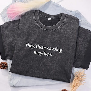 Peut inclure: Un t-shirt noir à l'aspect vieilli avec le texte "they/them causing may/hem" en blanc.