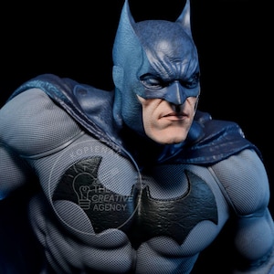 Puede incluir: Estatua detallada de Batman con un traje gris y azul, con el símbolo del murciélago en el pecho. La figura tiene una expresión seria y está sobre un fondo oscuro. El logotipo de The Creative Agency es visible.
