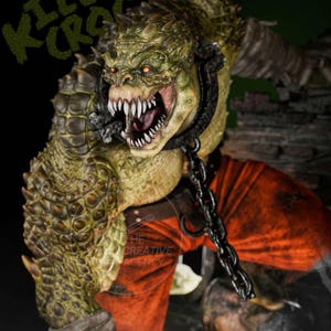 May include: A detailed sculpture of Killer Croc, a reptilian humanoid character. The figure has green and yellow textured skin, sharp teeth, and red eyes. It wears a metal collar and chain, with an orange shirt visible. The words "KILLER CROC" are in green text.
