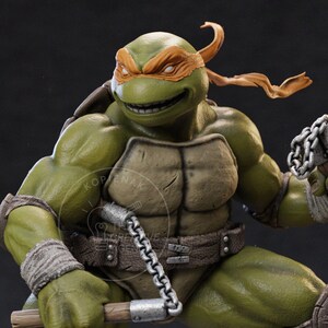 Può includere: Statuetta dettagliata di Michelangelo dei Teenage Mutant Ninja Turtles. La figura è verde con una maschera arancione, un guscio marrone e una cintura abbinata. Tiene in mano un'arma e ha un'espressione determinata.