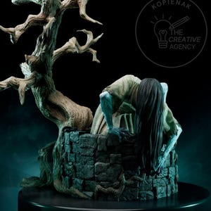 Puede incluir: Escultura detallada de una escena de terror. Una figura con largo cabello negro y tez pálida emerge de un pozo de piedra. Un árbol nudoso se alza detrás del pozo, aumentando la atmósfera inquietante. El logotipo de The Creative Agency es visible.