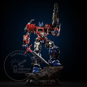 May include: A detailed red, blue, and silver Optimus Prime statue, holding a large gun and a sword. The figure is posed on a rocky base, with the logo of "The Creative Agency" in the lower left corner.
