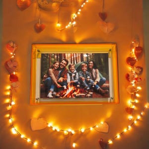 May include: A framed photo of a family of five enjoying a campfire in the woods. The photo is surrounded by string lights and hearts.