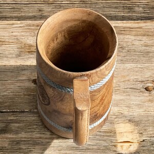 May include: A wooden beer mug with a handle. The mug is made of light brown wood and has two silver-colored rope bands around the body. The handle is also made of wood. The mug is sitting on a wooden surface.