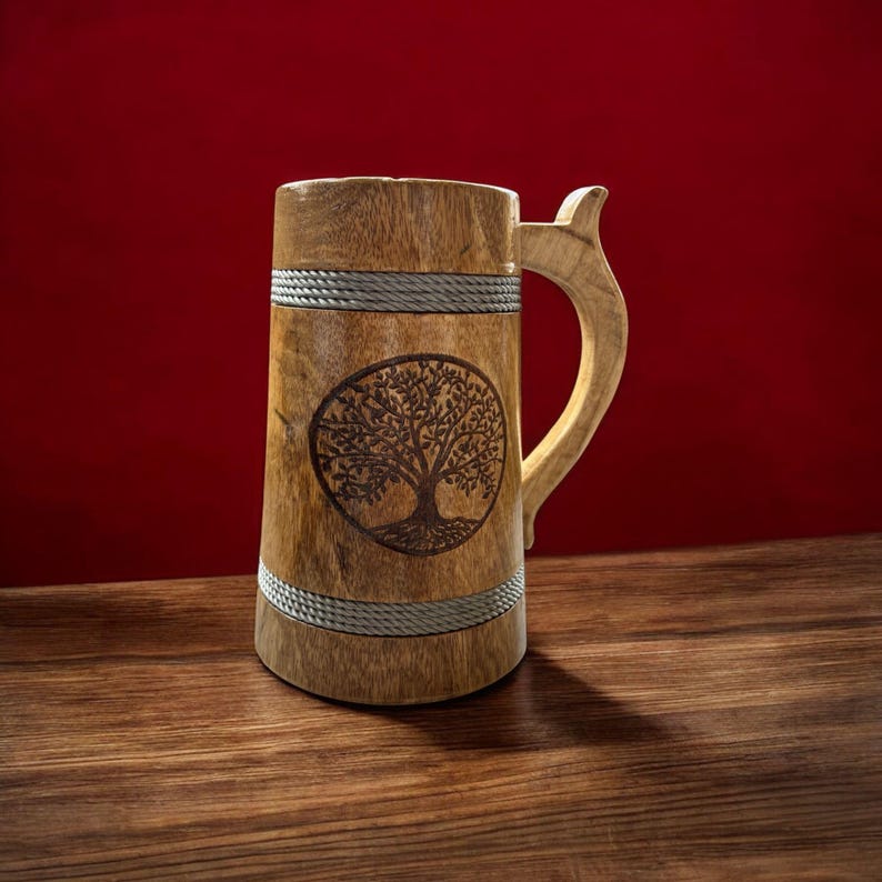 May include: Wooden beer mug with a carved tree design. The mug is made of light brown wood with a curved handle and a decorative rope detail. The tree design is inside an oval shape.