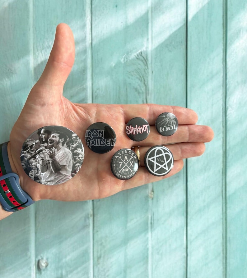 May include: A collection of seven round pinback buttons featuring band logos and images. The buttons are black and white, with the exception of one button that has a black background with a white and gray design. The buttons are held in a hand.