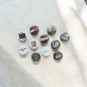 May include: A collection of round, black and white band button pins. The pins feature band names and logos, including "Black Flag," "Limp Bizkit," "Slipknot," and "Korn." The pins are arranged on a light-colored surface.
