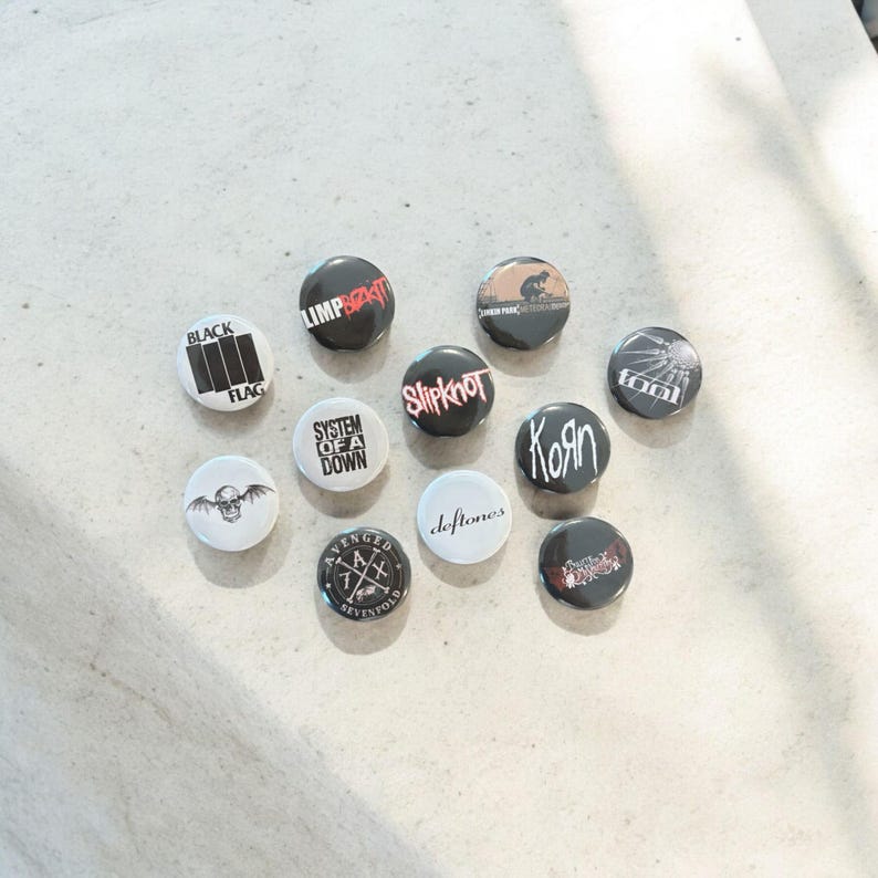 May include: A collection of round, black and white band button pins. The pins feature band names and logos, including "Black Flag," "Limp Bizkit," "Slipknot," and "Korn." The pins are arranged on a light-colored surface.