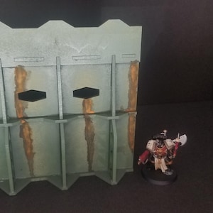 May include: A gray and brown textured wall with three rectangular openings. The wall is made of cardboard and is designed for use in tabletop games.
