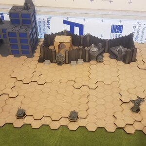Puede incluir: Un conjunto de terreno de juego de guerra de mesa con un campo de batalla modular de baldosas hexagonales. Incluye edificios en miniatura y fortificaciones en tonos grises y azules, junto con pequeños modelos de tanques. El terreno está sobre una superficie verde.