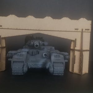 Könnte beinhalten: Ein graues Panzermodell mit einer großen Kanone, das durch einen hölzernen Torbogen fährt. Der Torbogen besteht aus mehreren Holzstücken und hat ein schlichtes Design.