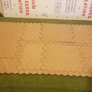 May include: A collection of interlocking, light brown, hexagonal tiles arranged on a green surface. The tiles are made of wood and are designed to fit together, forming a larger surface. The image is taken from a top-down perspective.