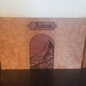 May include: A three-panel wooden game board with a mountain scene and the name "Andrew" engraved on the center panel. The panels are decorated with a wood grain pattern.