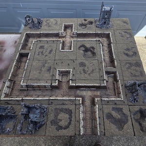 May include: A detailed tabletop gaming terrain set. The modular design features a central pathway with stone walls and various structures, including a tower and ruins. The color palette is primarily gray and brown, creating a realistic, weathered appearance. The set is designed for role-playing games.
