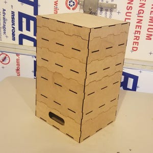 May include: A brown wooden box with a decorative design. The box has a series of slots cut into the front and sides. The box is made of plywood and has a natural wood finish.