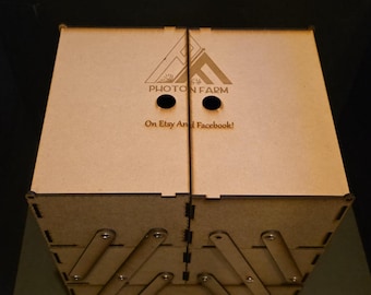 May include: A light brown wooden box with a hinged lid and a multi-tiered, expanding design. The box features the text "PHOTON FARM" and "On Etsy And Facebook!" on the lid. The box has two circular cutouts and a geometric design.
