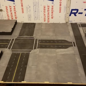 以下が含まれることがあります： テーブルゲーム用のモジュール式のグレーと黒の地形セット。セットには、黄と白の線が入った道路セクション、灰色の歩道、建物のプラットフォームが含まれています。木製の表面に配置されています。