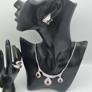 Könnte beinhalten: Ein Schmuckset bestehend aus einer silberfarbenen Halskette, Ohrringen und einem Ring, verziert mit tropfenförmigen roten Steinen und kleinen klaren Kristallen. Die Kette hat drei Anhänger, während der Ring zwei rote Steine zeigt.