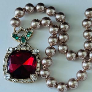 May include: A necklace featuring a strand of metallic bronze-toned pearls and a large square pendant. The pendant has a deep red faceted center stone framed by clear crystals and a top accent piece set with small green rectangular stones.
