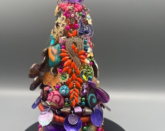 May include: A decorative, cone-shaped object adorned with a variety of colorful embellishments. The base is a deep magenta, supporting layers of beads, gems, and trinkets in shades of purple, orange, and teal. A large, faceted purple gem tops the piece.