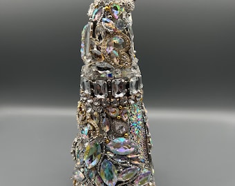 Puede incluir: Una botella decorativa con un tapón de cristal, profusamente adornada con piedras preciosas iridiscentes y transparentes. La botella está adornada con cristales de varias formas y tamaños, creando una apariencia brillante y lujosa. La base es plateada.