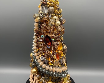 May include: A decorative, handcrafted tree-shaped object. It is adorned with a variety of beads, gems, and embellishments in shades of gold, brown, and amber. The tree is topped with a yellow crystal. The base is a dark, circular platform.