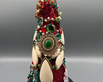 May include: A decorative bottle with a faceted green stopper. The bottle is covered in red sequins and adorned with green, red, and white jewels, pearls, and gold accents. Cream-colored, teardrop-shaped embellishments are also present.