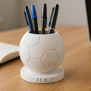 Peut inclure: Un pot à crayons blanc en forme de ballon de football avec le nom "ALEX" sur la base. Le pot contient plusieurs stylos, dont un bleu. Le pot à crayons est posé sur un bureau en bois à côté d'un cahier ouvert et d'un ordinateur portable.