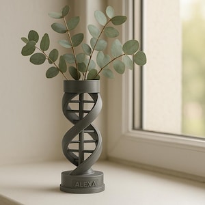 Puede incluir: Un jarrón gris con diseño de hélice de ADN, con ramas de eucalipto verdes. El jarrón tiene una base cilíndrica con el nombre "ALEXA" inscrito. El jarrón está colocado en un alféizar blanco cerca de una ventana.