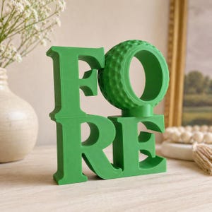 May include: A green decorative piece spelling "FORE" with a golf ball design for the "O". The letters are in a bold, block style. The piece is made of a solid material and sits on a light-colored surface.