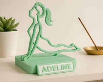Peut inclure: Figurine de yoga vert menthe représentant une personne en posture du chien tête en haut, avec le nom "ADELINE" sur la base. Un petit cactus en pot blanc est à gauche.