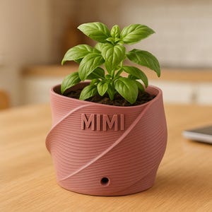 Puede incluir: Una planta de albahaca en una maceta rosa texturizada con el nombre "MIMI" en relieve. La maceta tiene un diseño en espiral y un orificio de drenaje. La planta está sobre una mesa de madera.