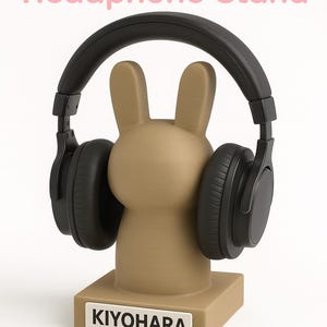 May include: A tan bunny-shaped headphone stand with black headphones. The stand has a rectangular base with the text "KIYOHARA" in a white rectangle. The words "Bunny Headphone Stand" are in pink text above the stand.