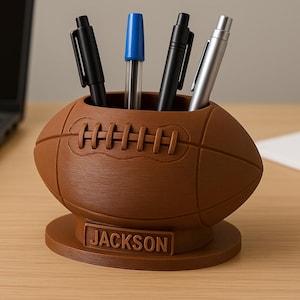 May include: Brown football-shaped pen holder with the name "JACKSON" on the base. The holder contains four pens: two black, one blue, and one silver. The pen holder is on a wooden desk.