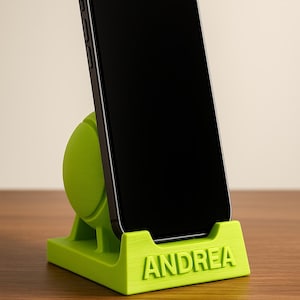 May include: A lime green phone stand with the name "ANDREA" printed on the front. A black smartphone is placed in the stand, displaying a black screen. The stand has a rounded back and a rectangular base.