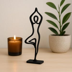 May include: Black metal sculpture of a person in a yoga pose, standing on a black base. The sculpture is on a wooden surface with a candle and a potted plant in the background.
