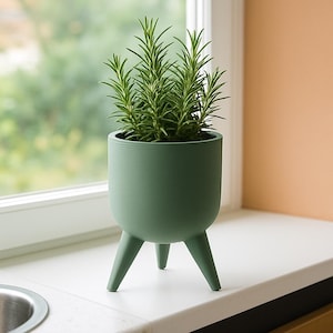 May include: A green, three-legged planter pot with a rosemary plant inside. The pot is a muted green color and has a rounded shape. The rosemary plant has long, thin, green leaves. The planter is sitting on a white windowsill.